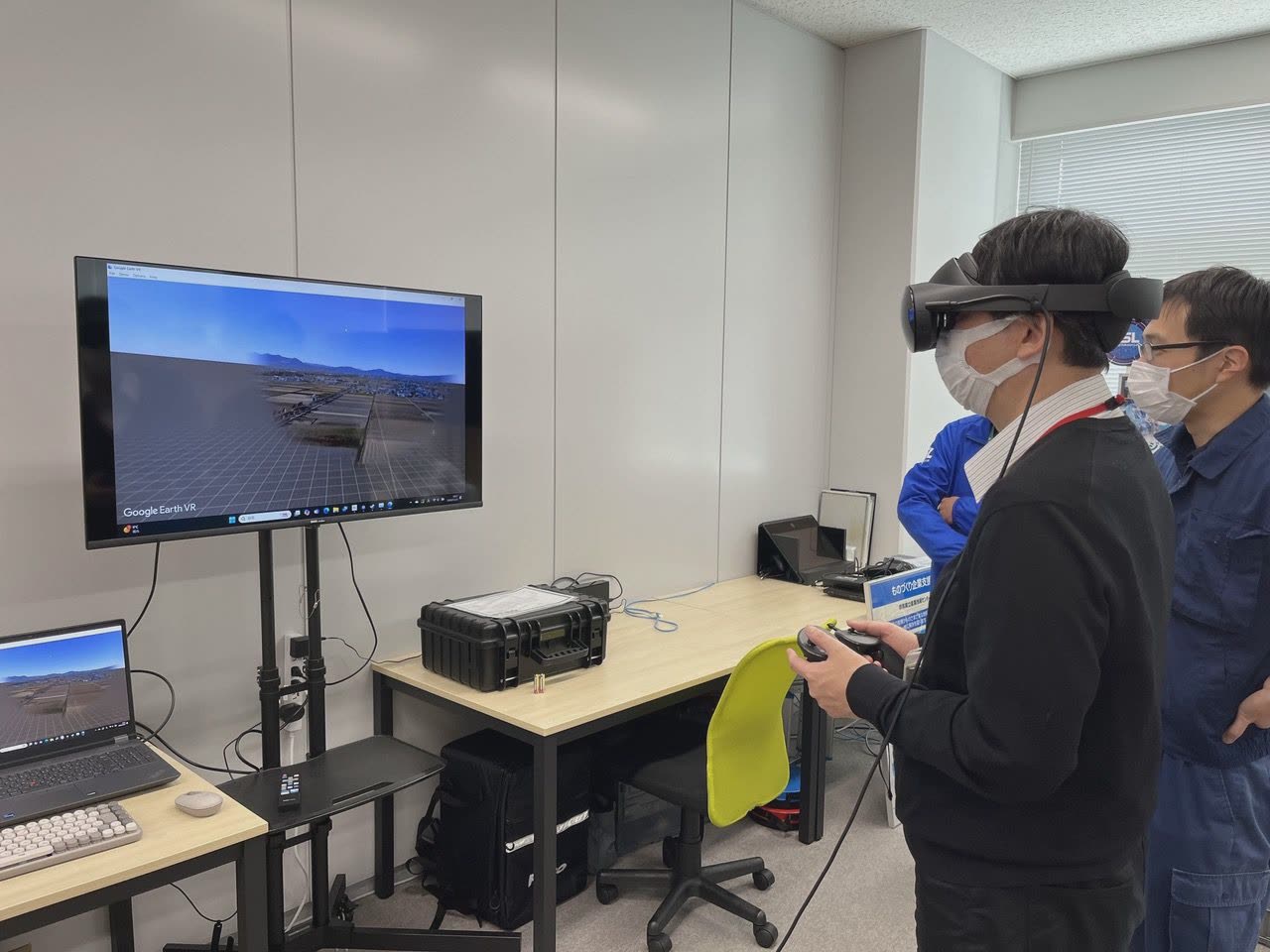 VRゴーグルを着けた男性がコントローラーを持ち、横のモニターに Google Earth VR の景色が映っている様子