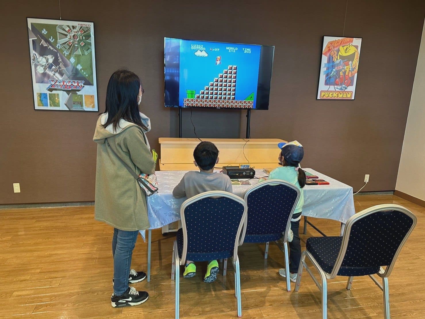 壁の大型モニターにスーパーマリオブラザーズを映し、テーブルで子ども2人がファミコンを遊び、大人が後ろから見守っている様子